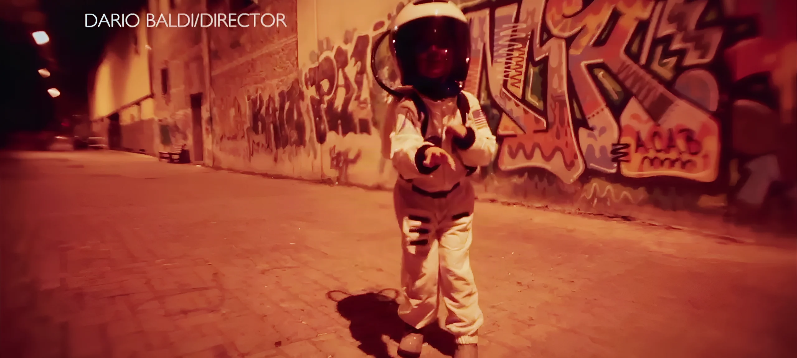 Un astronauta in tuta chiara indica la camera in un vicolo notturno, davanti a un muro coperto di graffiti.